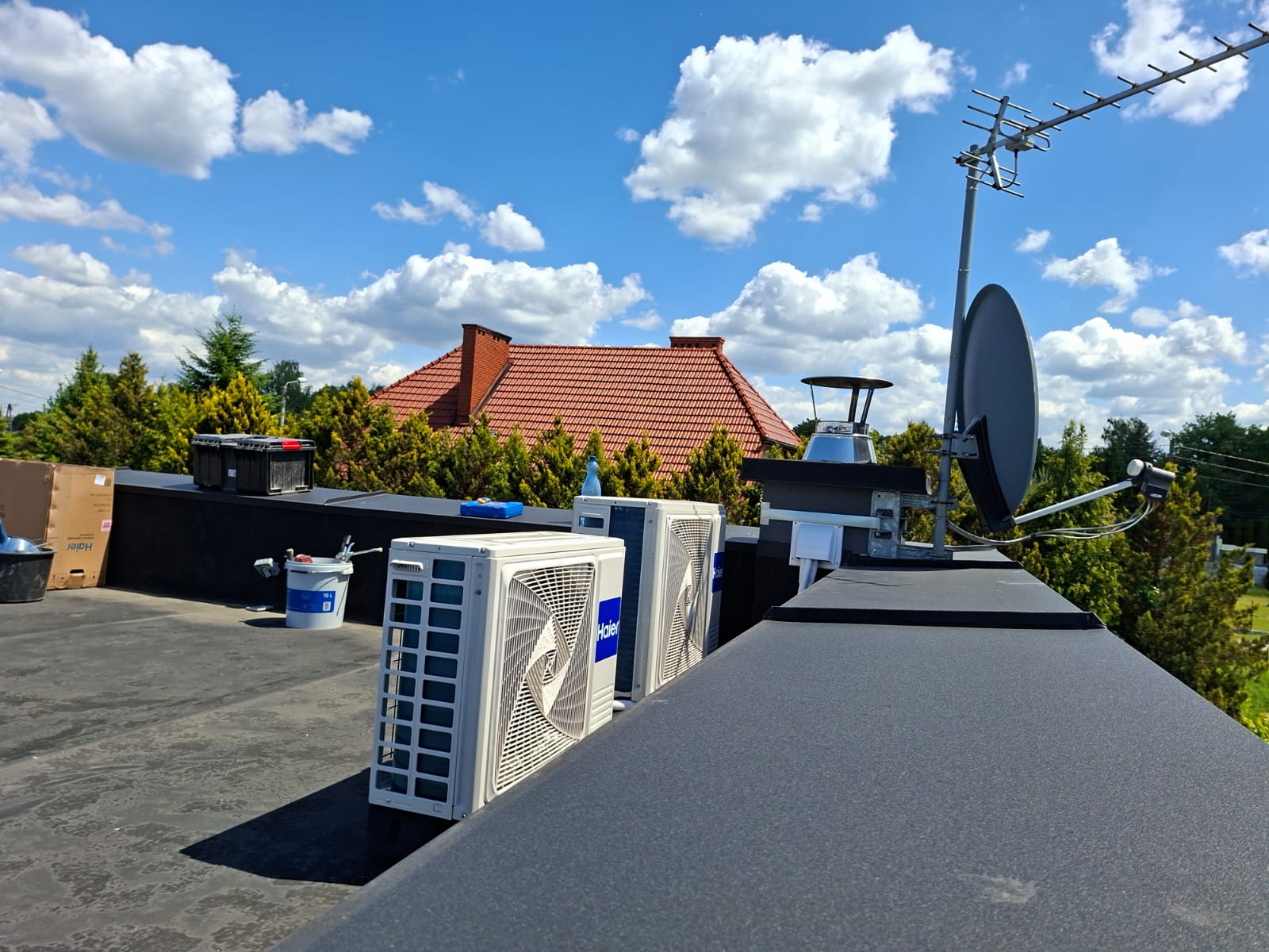 Ekipa Alfaprojekt na dachu podczas instalacji dwóch jednostek zewnętrznych pomp ciepła Haier. Obok widać narzędzia, materiały i antenę satelitarną na tle nieba.
